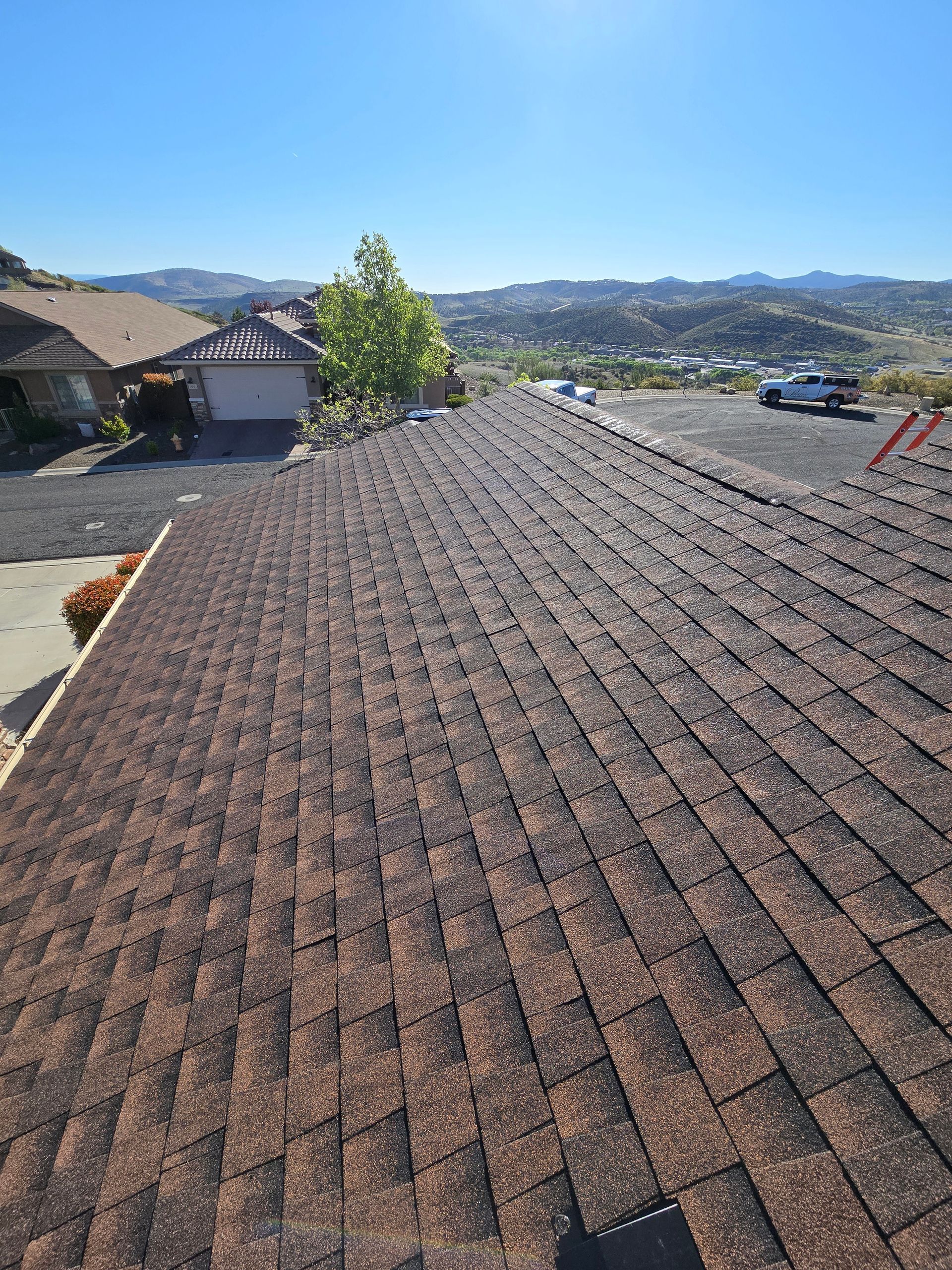 Rooftop with brown shingles overlooking a suburban neighborhood and distant mountains under a clear blue sky