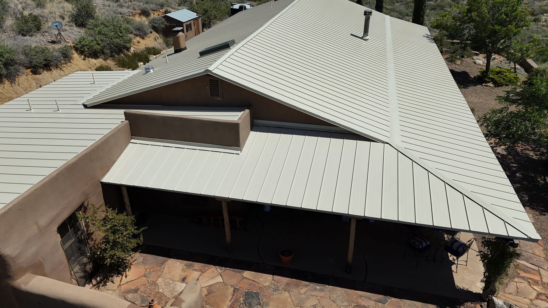 Aerial view of a light-gray metal-roof house surrounded by trees and rocky ground.