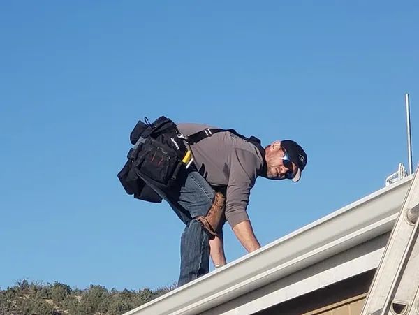 Person on a roof, leaning over a gutter, wearing a tool belt and sunglasses against a blue sky.