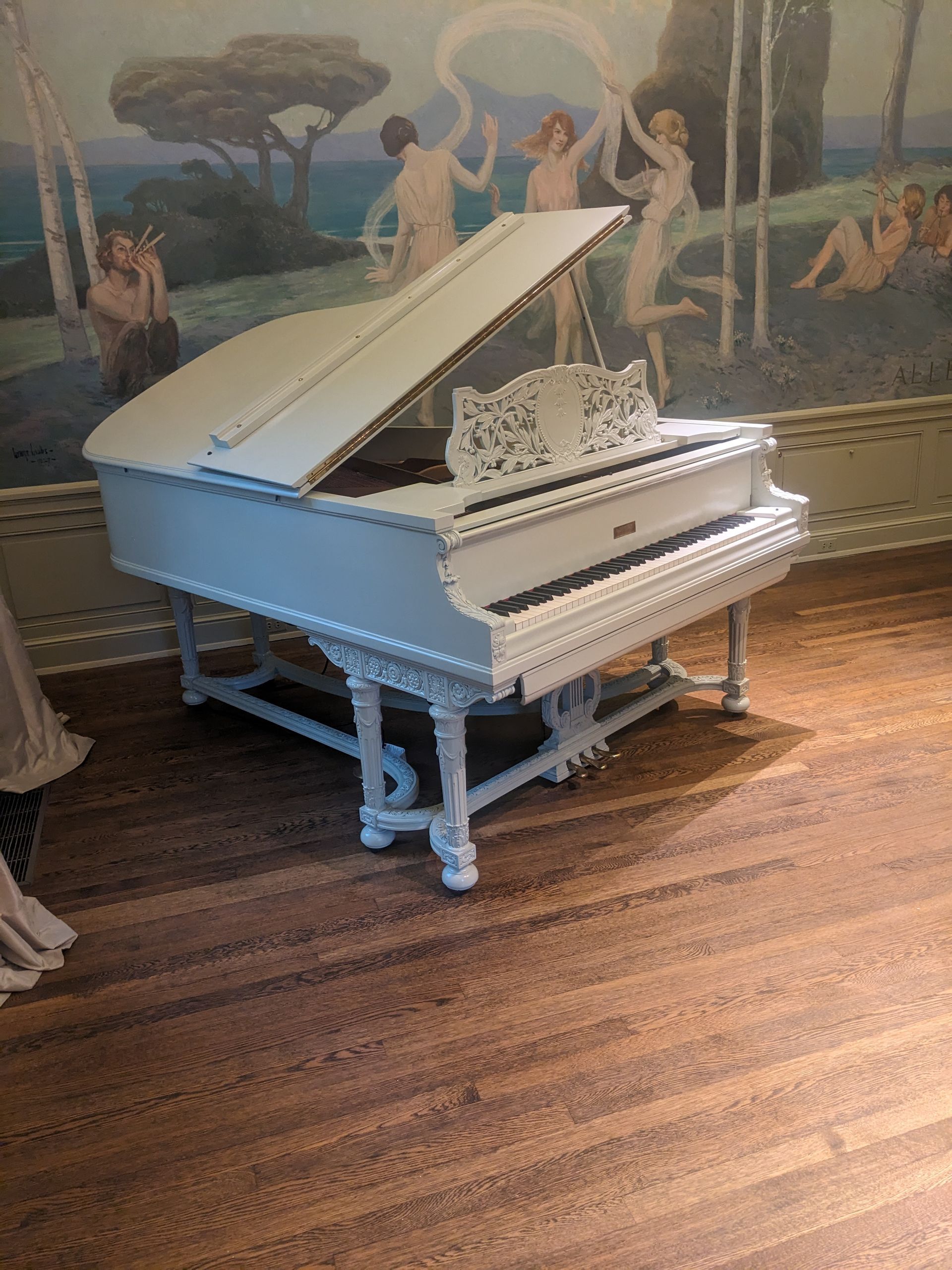 A white grand piano is sitting on a wooden floor in front of a painting.