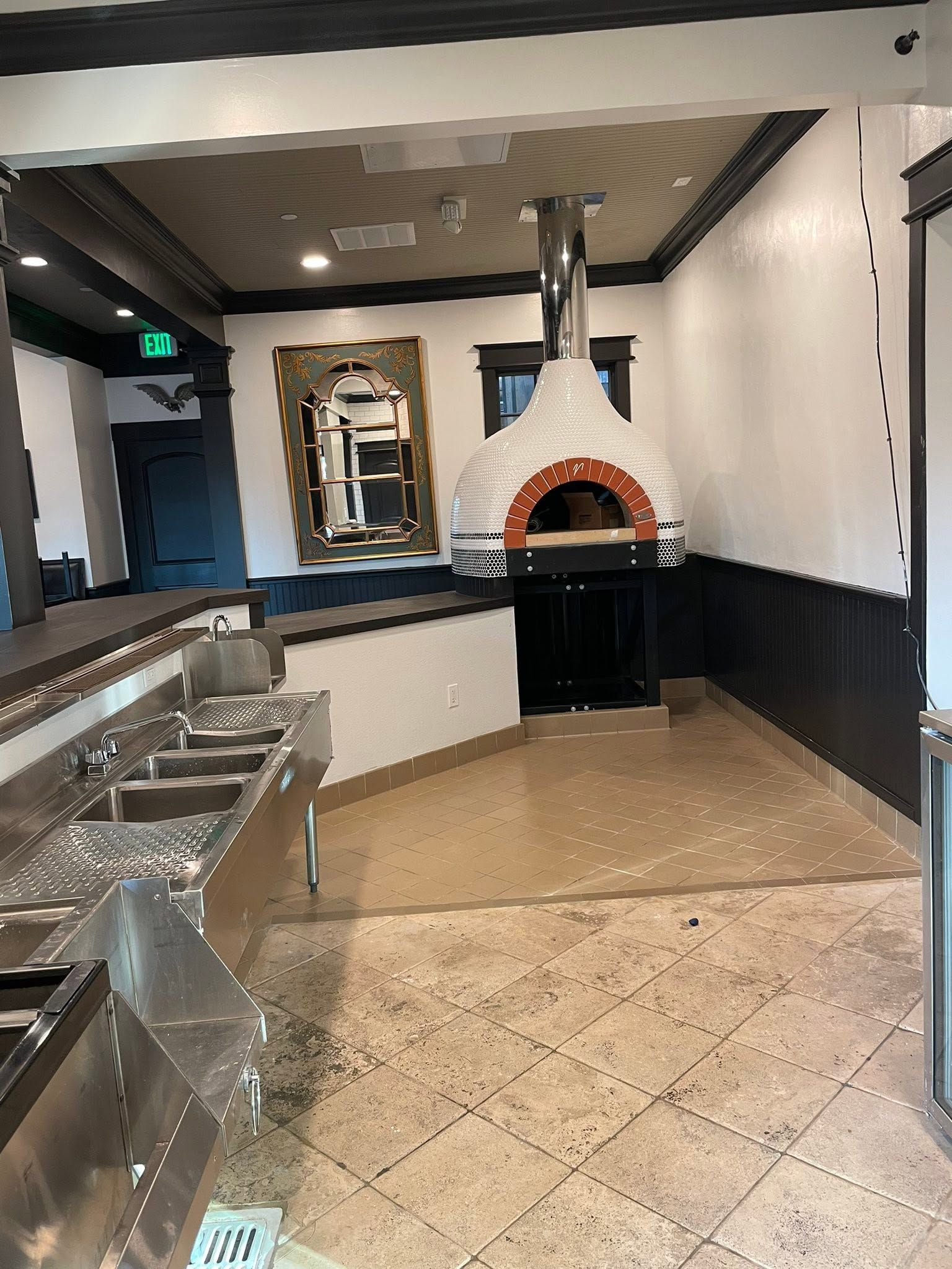 Empty restaurant bar area with a tiled floor, stainless counter, and wood-fired oven against the back wall