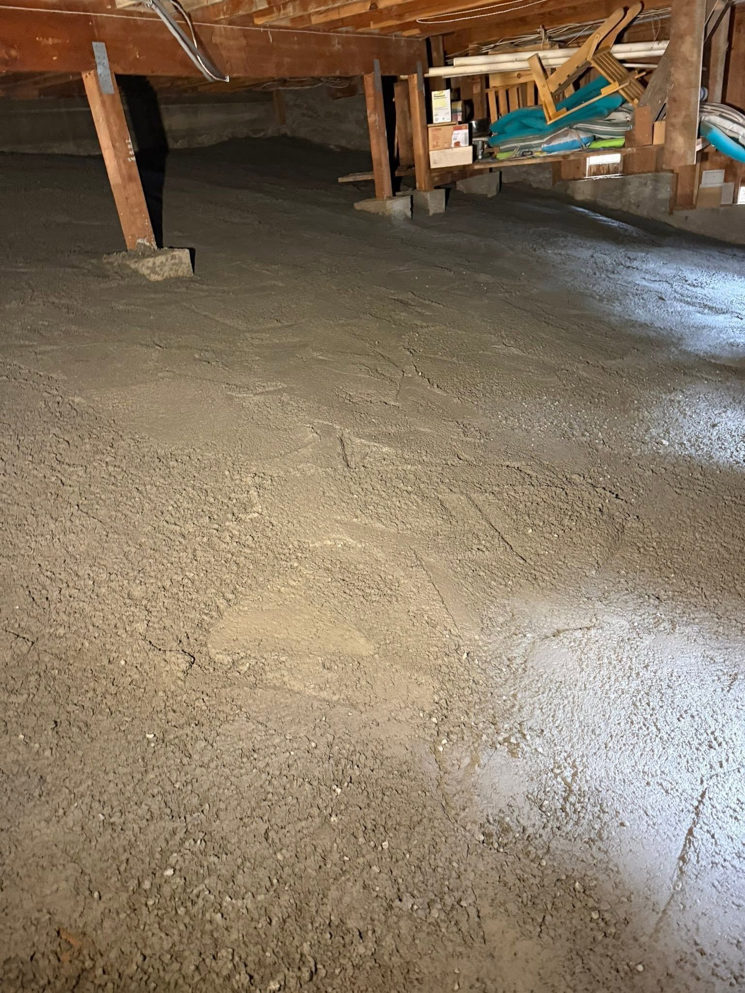 Under a wooden floor, a concrete crawlspace with support posts and stored items in the background.
