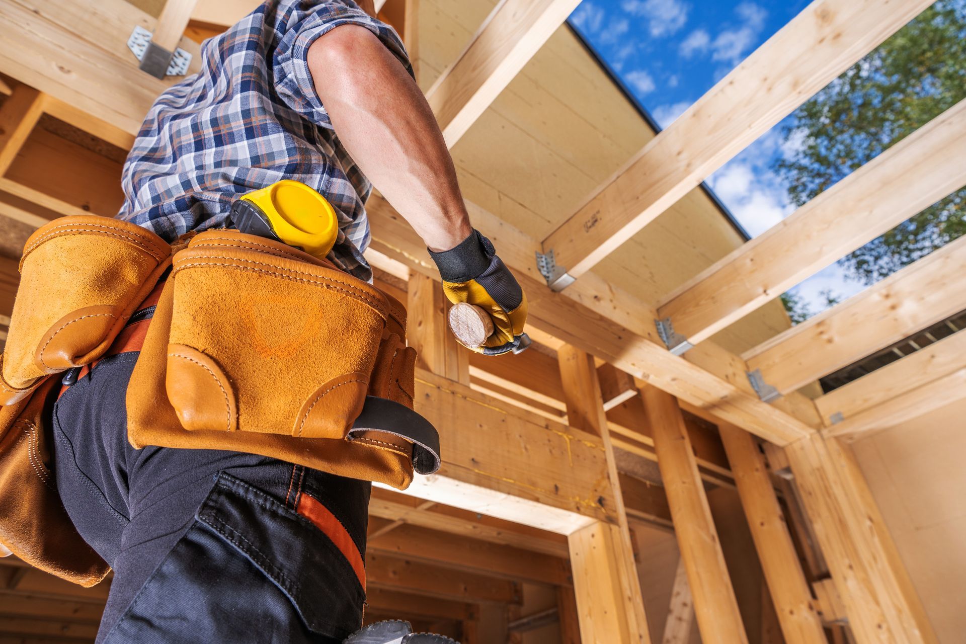 Construction worker installing ceiling beams in a wooden house frame, wearing a tool belt and gloves