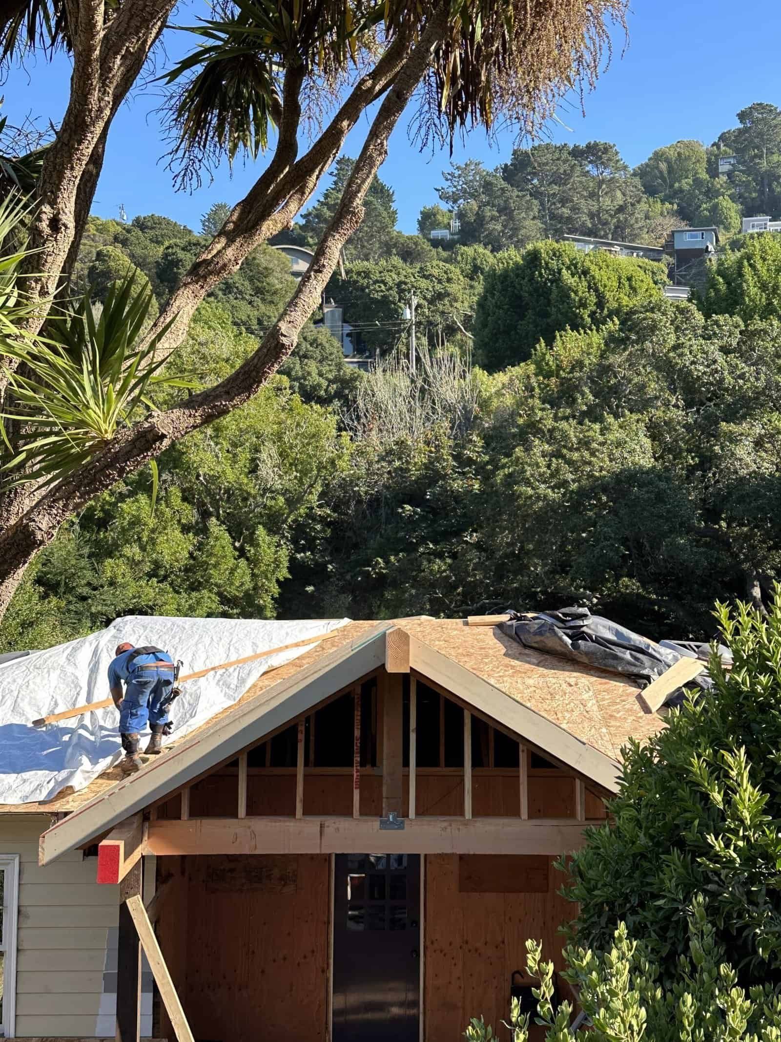 Worker on a roof of a brick house surrounded by dense green trees and hillside vegetation.