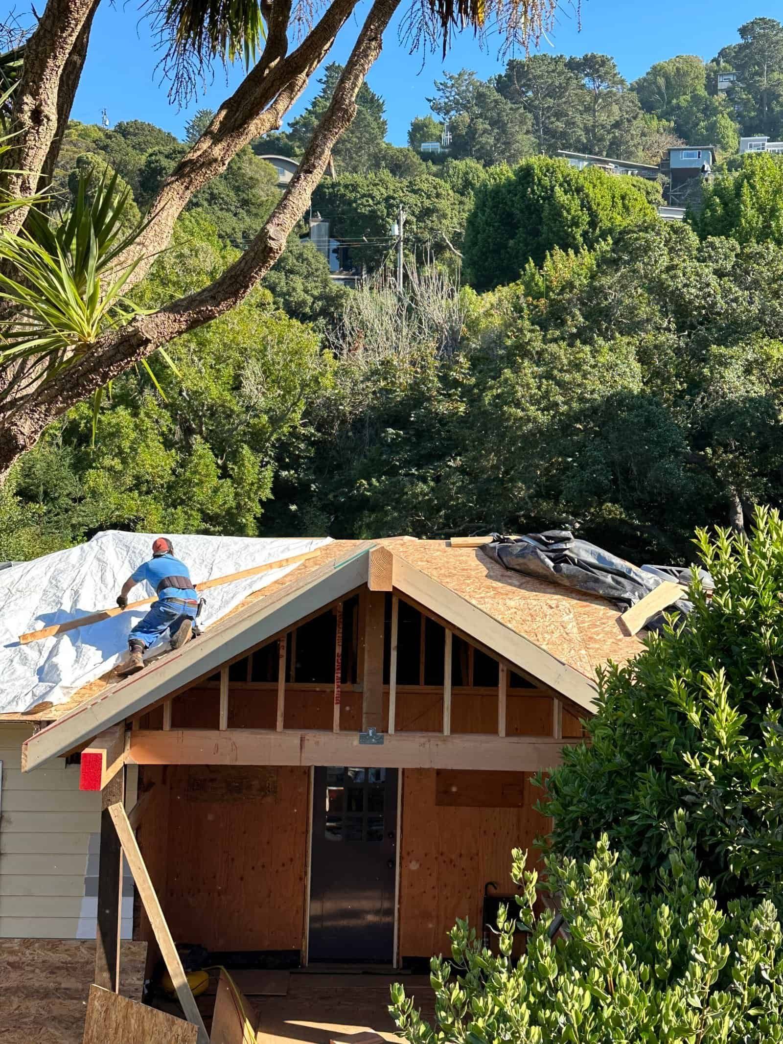 Workers repairing a small house roof amid lush green trees, with one worker on the roof.