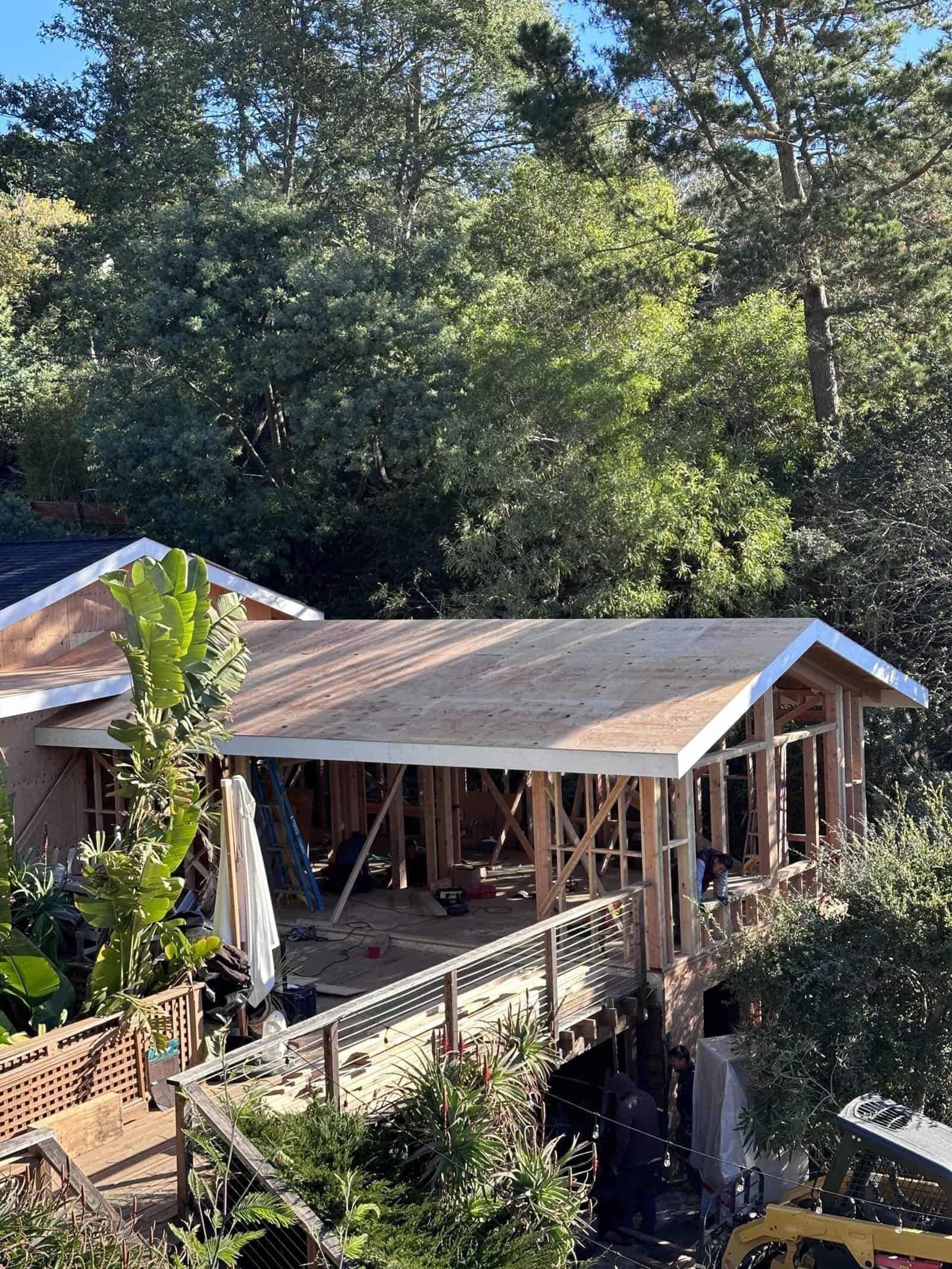 Wooden pavilion under construction in a wooded area, with a sloped roof and open framing.