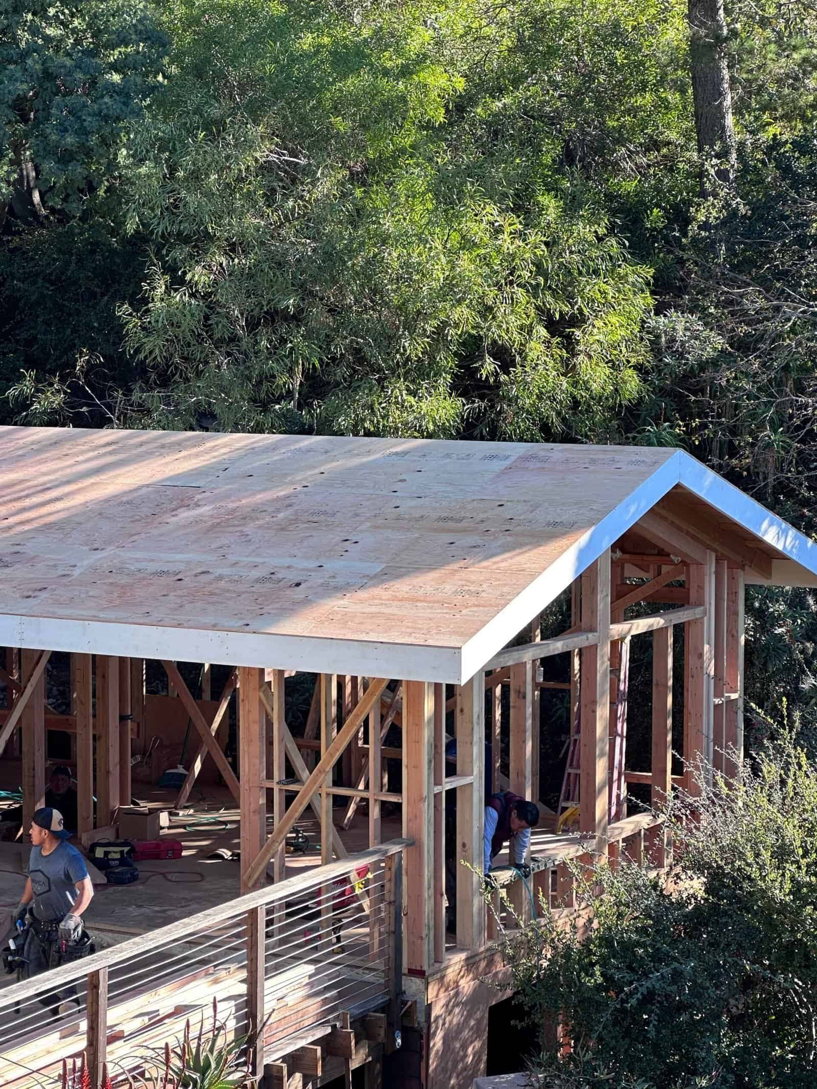 Partially built wooden structure with a tan roof, surrounded by trees and fencing outdoors