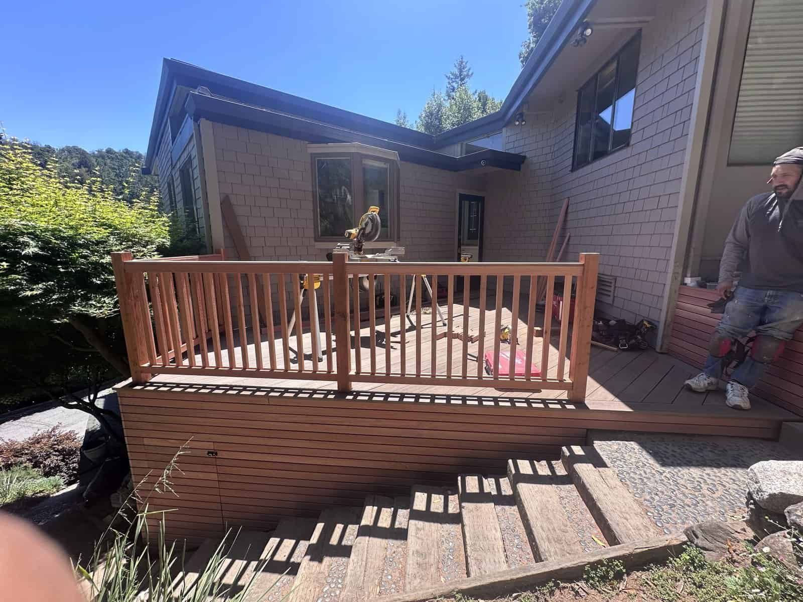 Wooden deck with railing beside a brick house, with a person standing at the right edge.