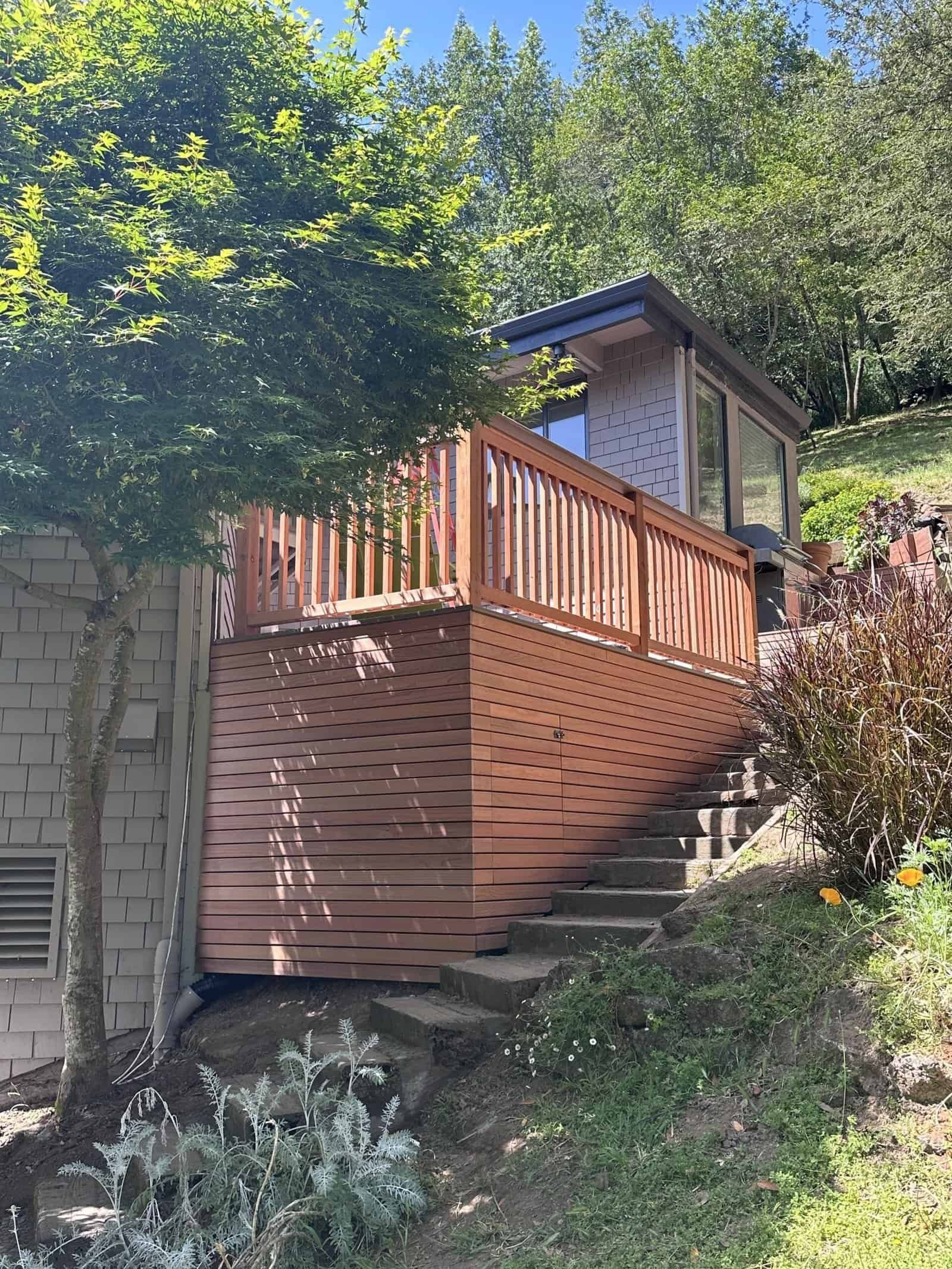 Wooden deck stairs and railing on a hillside beside a house, surrounded by trees and greenery.