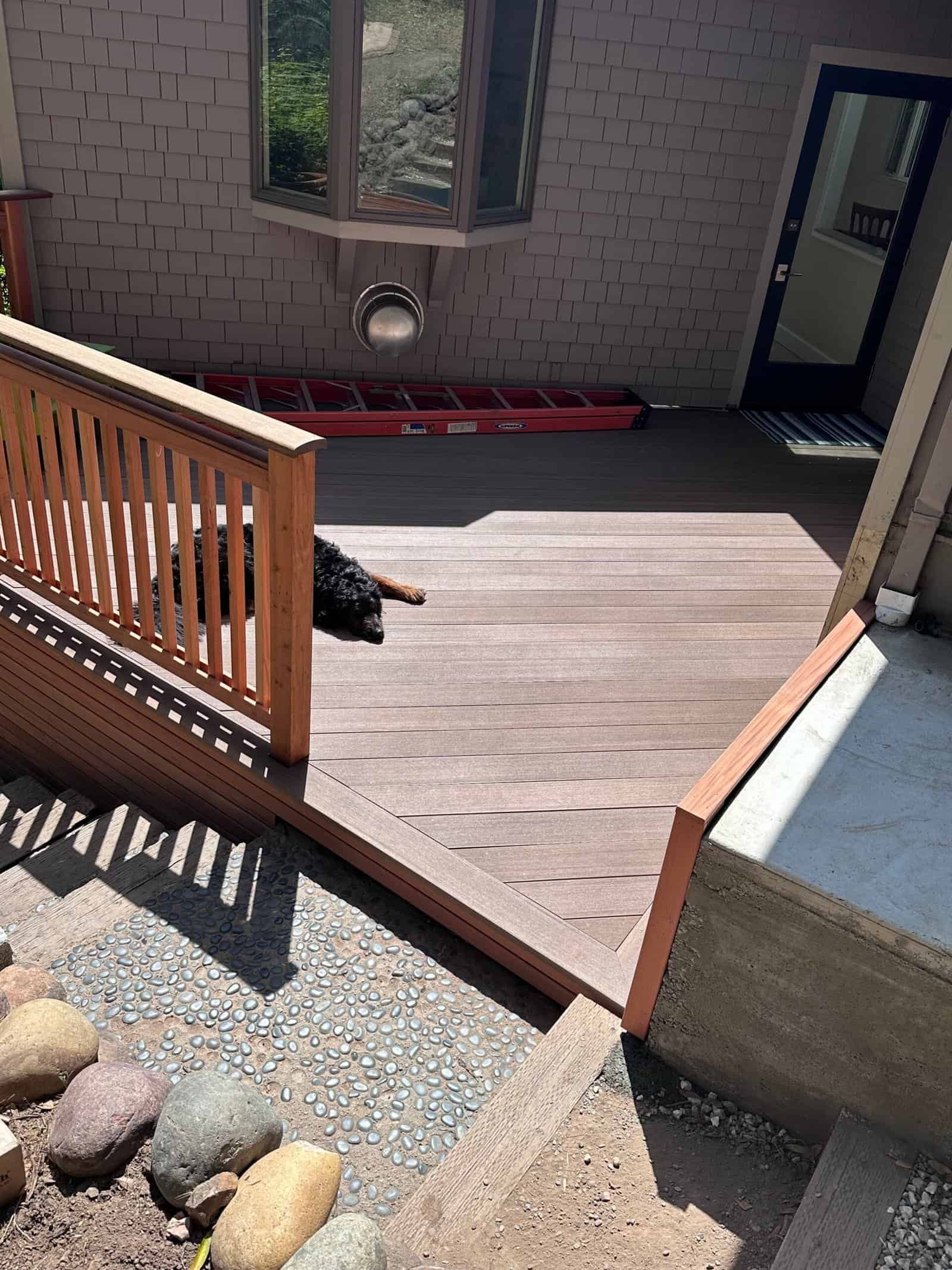 Wooden deck with railing and open doorway beside a brick house, with a small black dog lying in the sun.