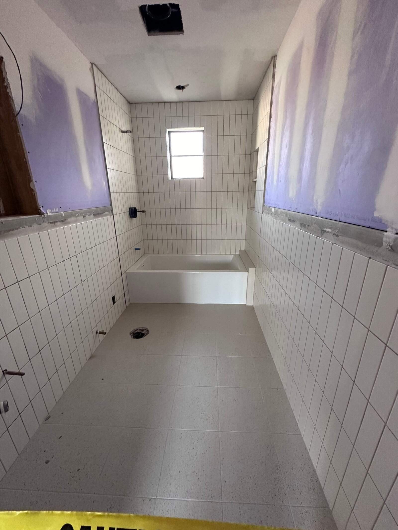 Narrow white-tiled bathroom under renovation with a small window and unfinished tub area.