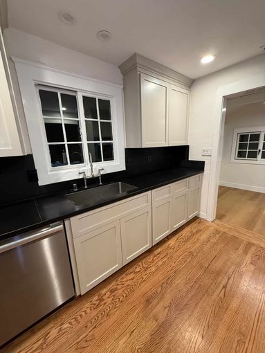 Bright kitchen with white cabinets, black countertops, stainless dishwasher, and hardwood floor