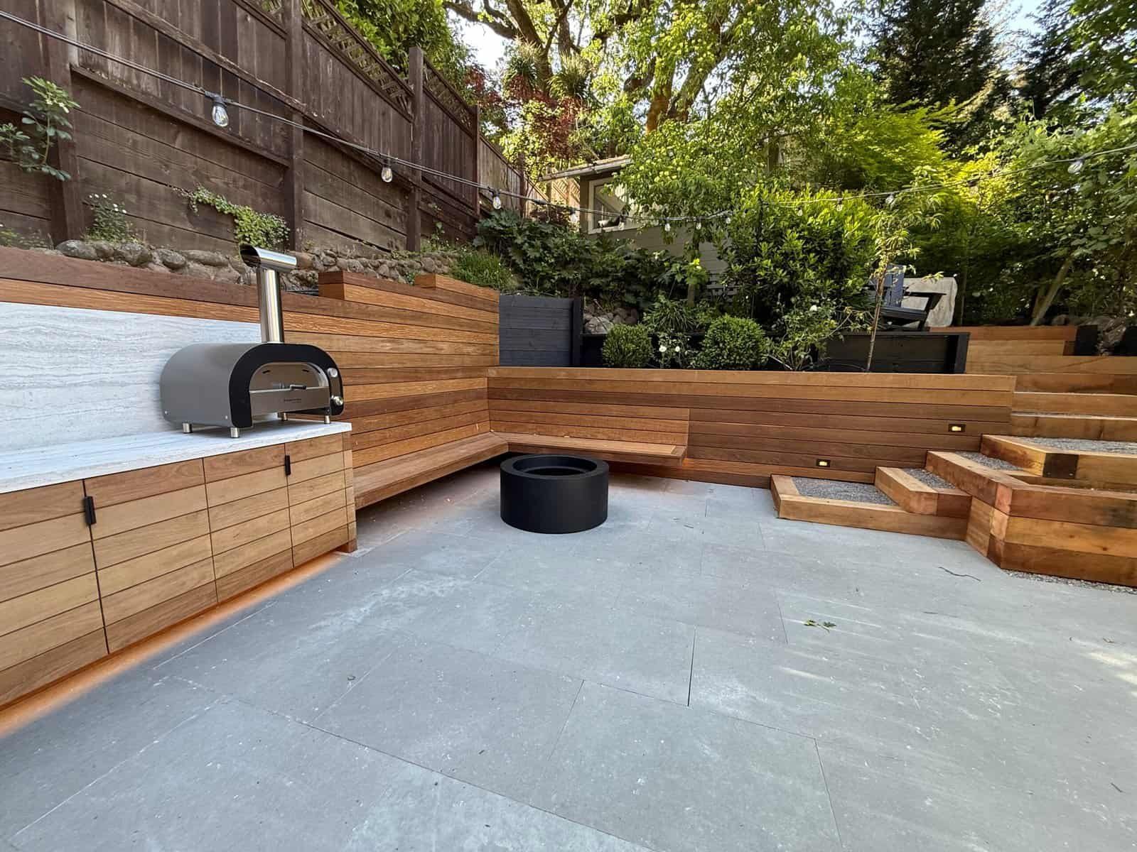 Backyard patio with wooden benches, concrete floor, brick wall, and trees on a terraced slope