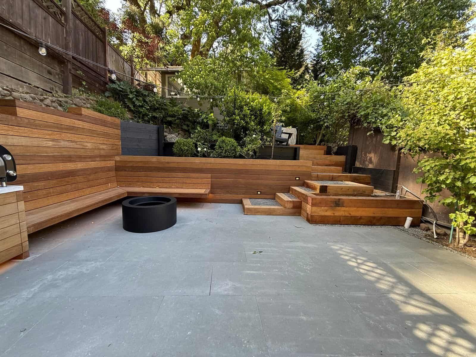 Backyard patio with wooden seating, a black fire pit, and trees casting dappled shade