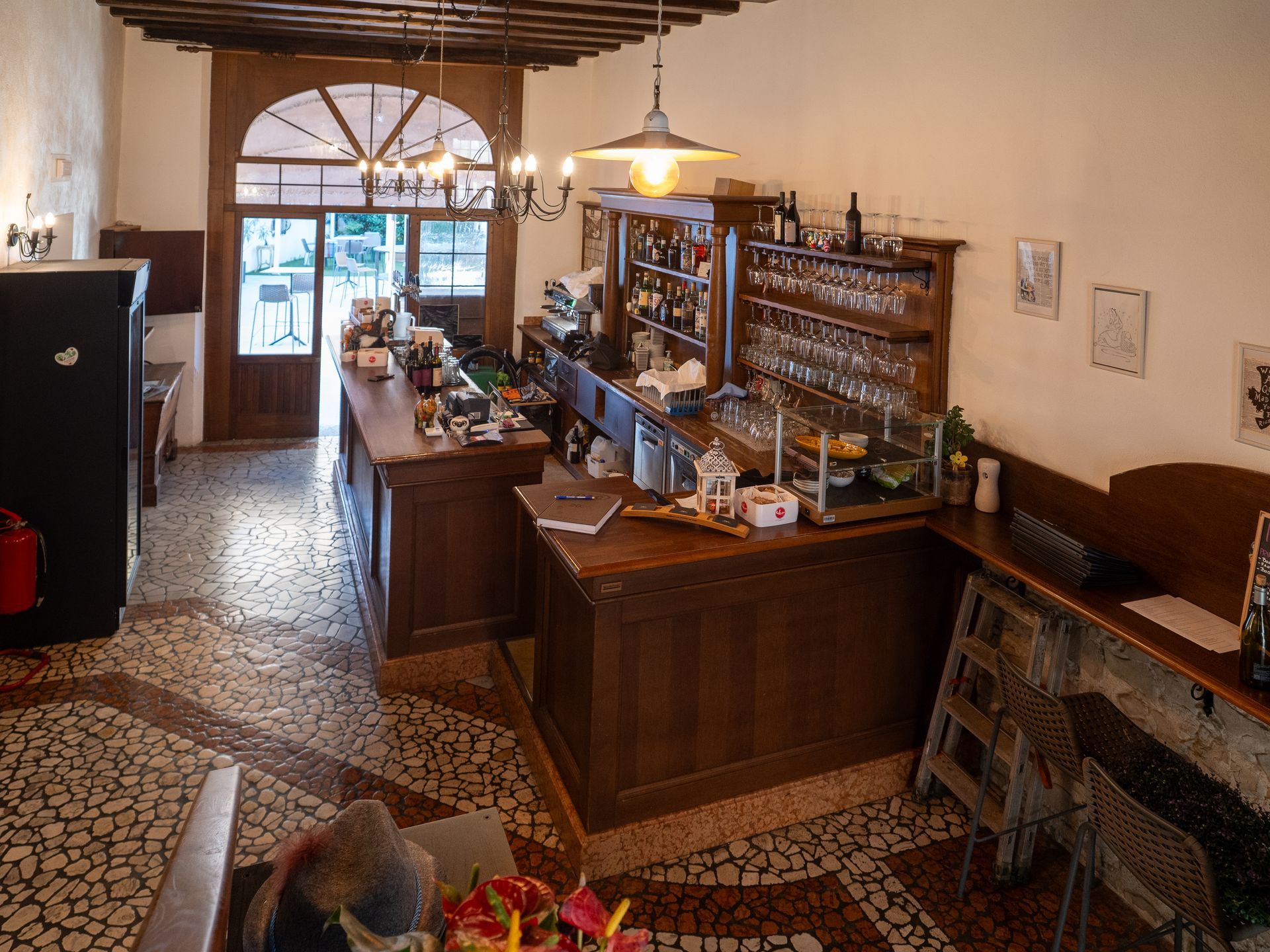 Vista interna di un bar rustico con bancone in legno, scaffali pieni di bicchieri e un ingresso verso l'esterno.