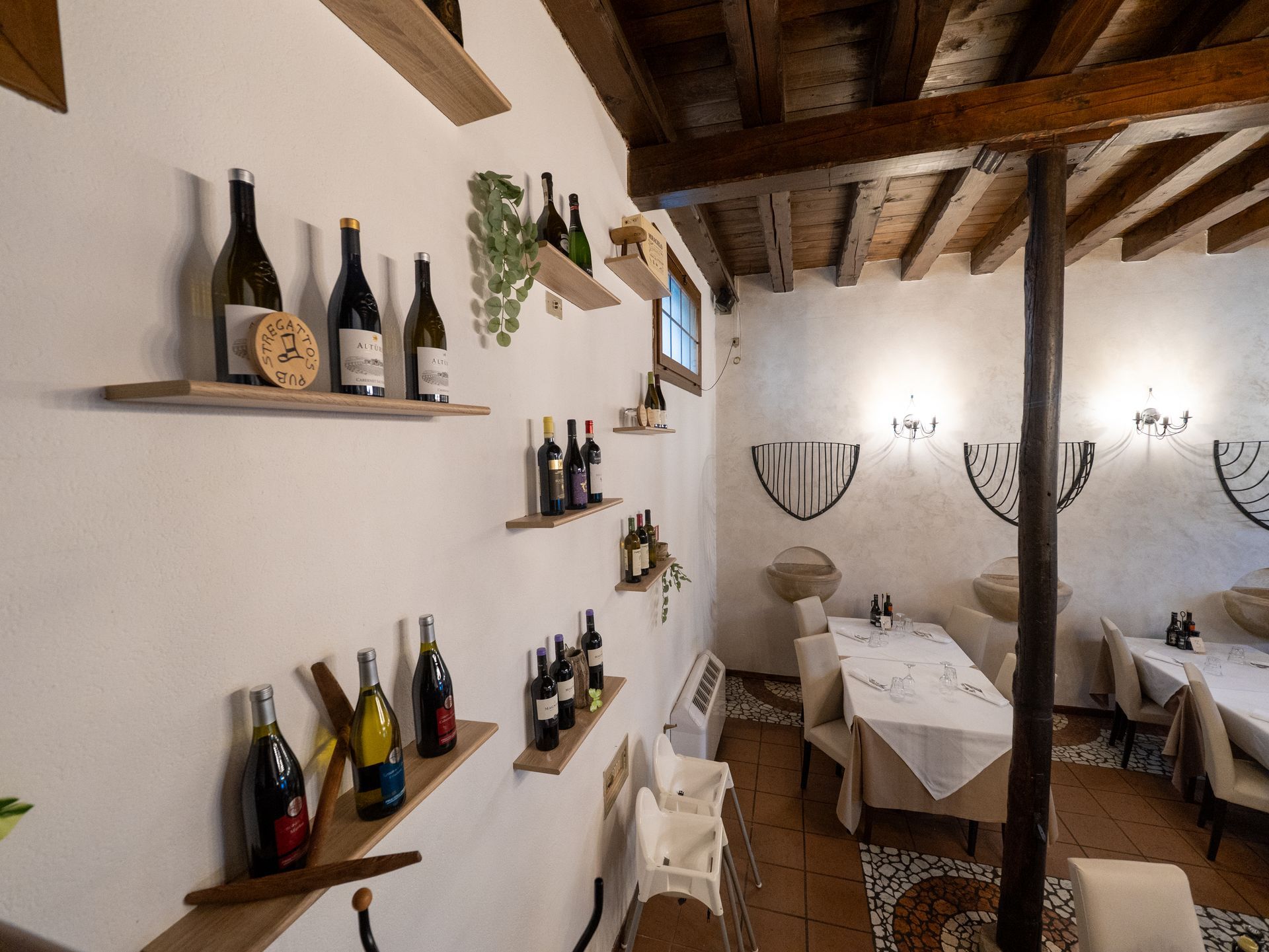 Interno di un ristorante con bottiglie di vino sugli scaffali, tavoli apparecchiati con tovaglie bianche e soffitto con travi in legno.