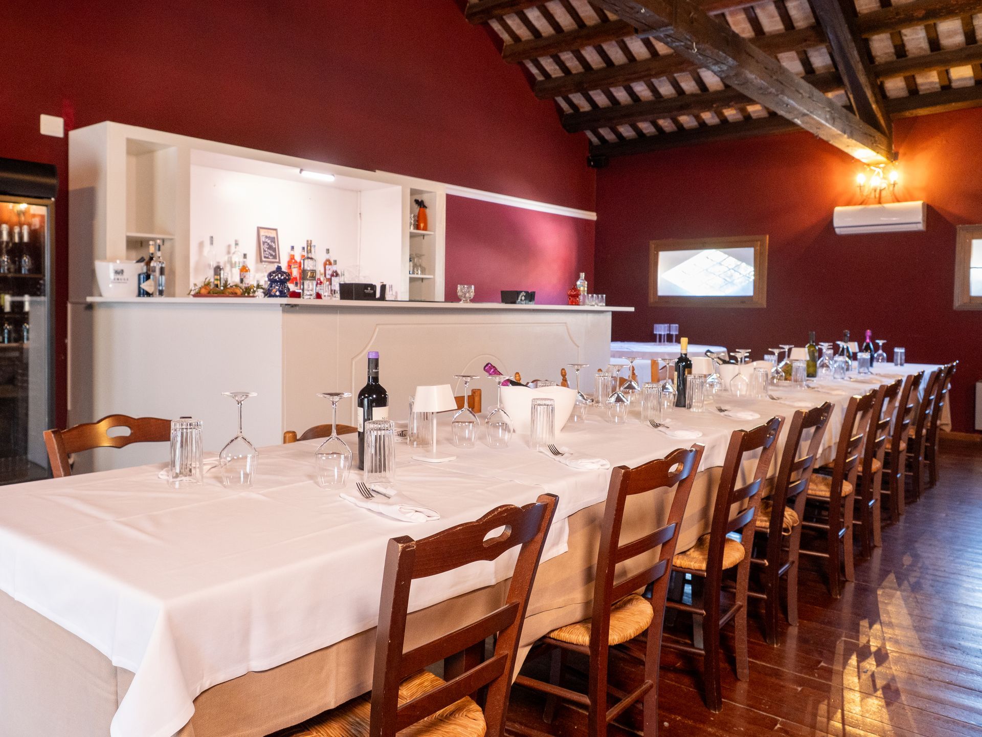 Sala da pranzo con lungo tavolo apparecchiato per un pasto, sedie, zona bar sullo sfondo, pareti color marrone.