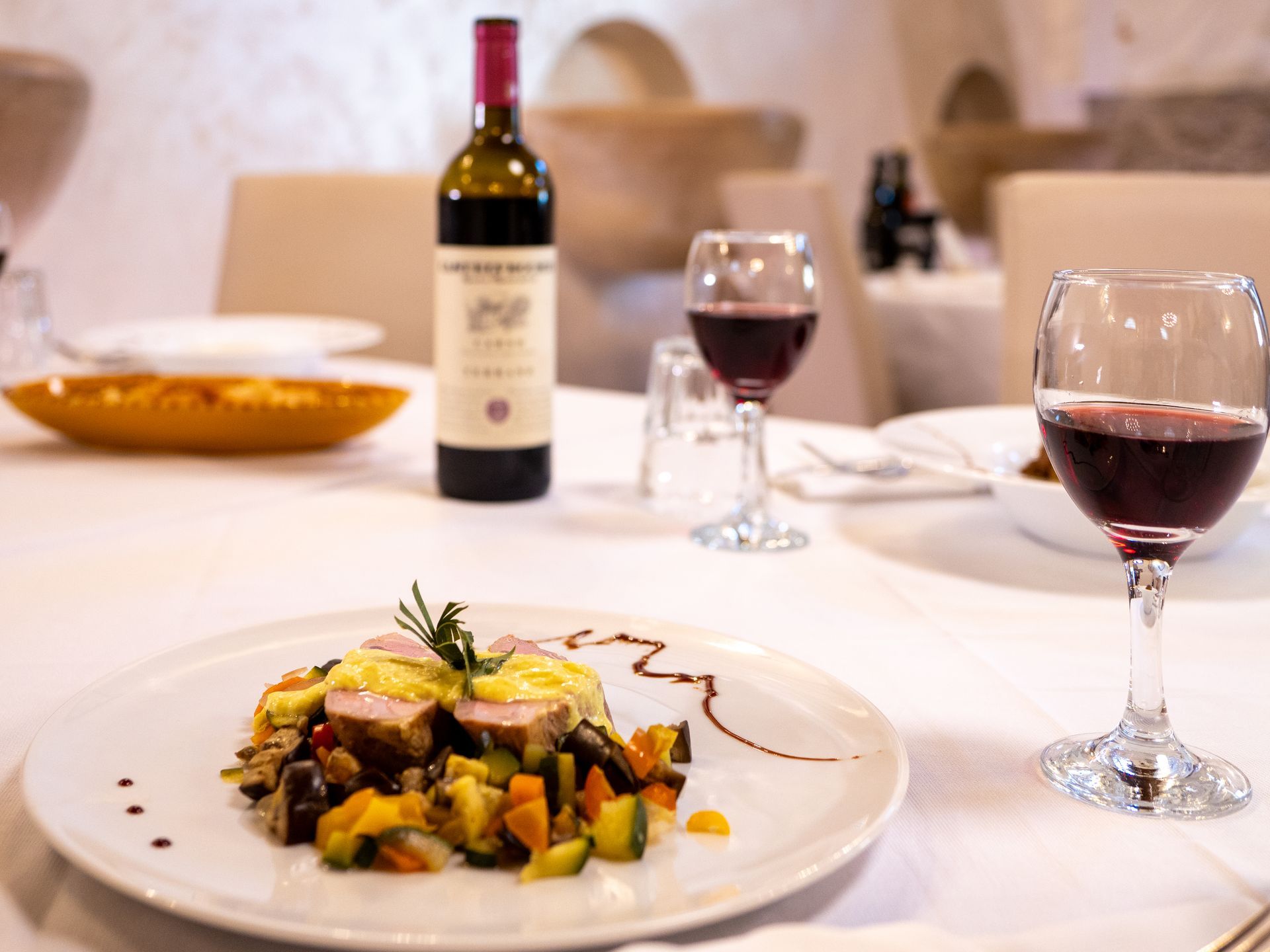 Piatto di cibo con verdure, bicchieri di vino e una bottiglia di vino rosso su una tovaglia bianca.