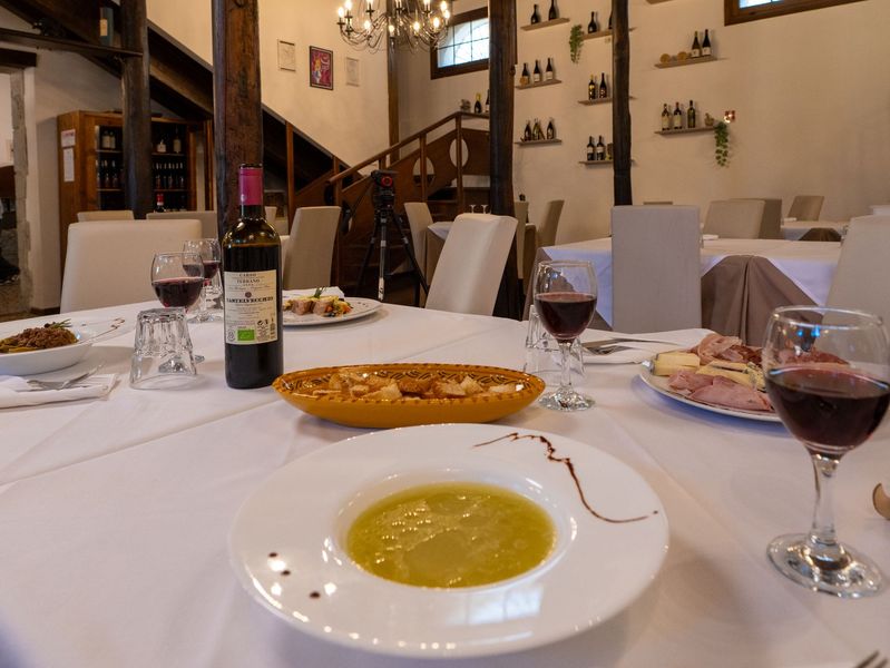 Tavolo del ristorante apparecchiato con piatti, vino e una bottiglia di vino. Stanza dai colori chiari, travi in legno e scale sullo sfondo.