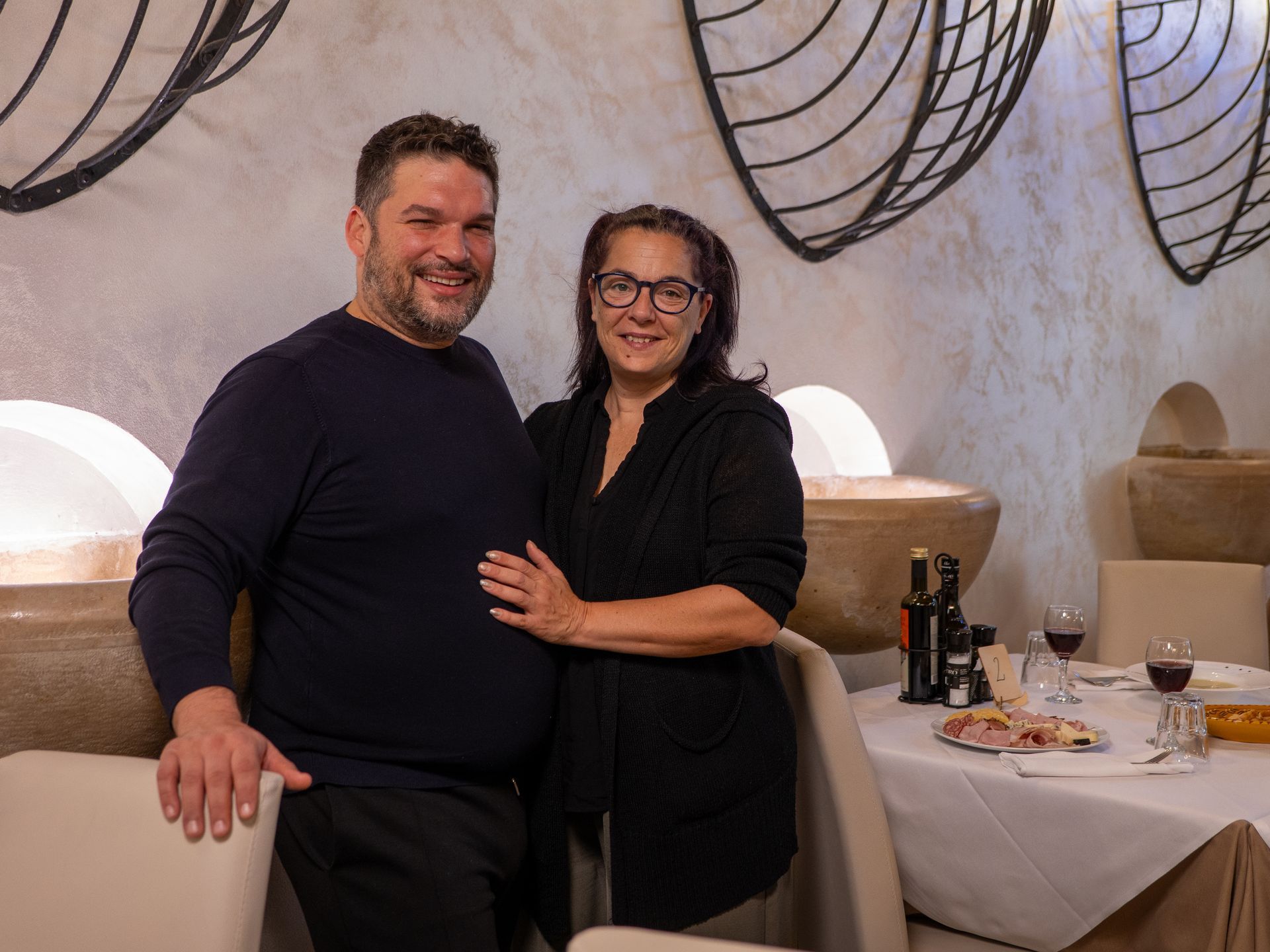 Coppia sorridente al ristorante, in piedi accanto a un tavolo apparecchiato per la cena.