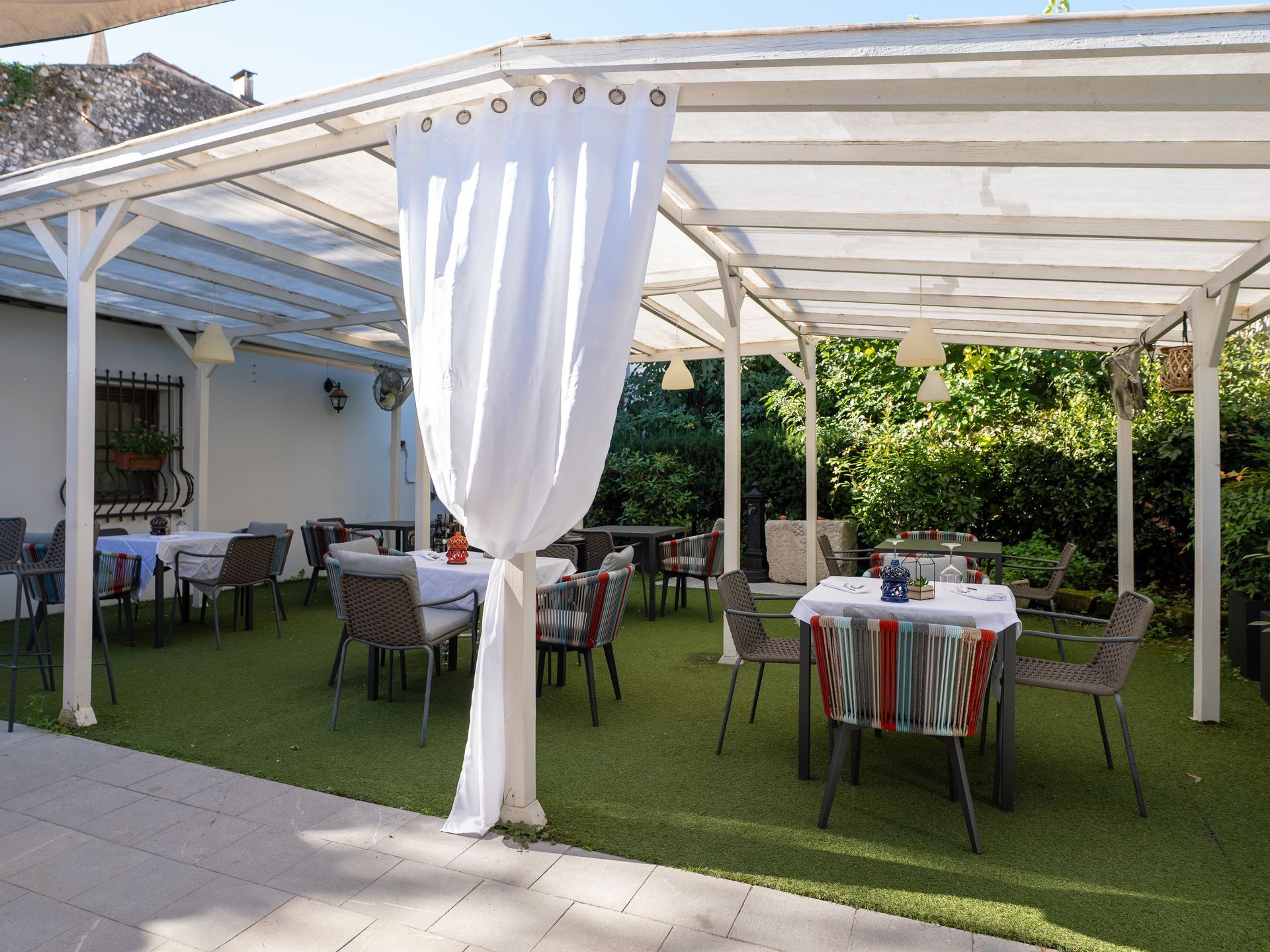Area esterna del ristorante con posti a sedere sotto una tettoia bianca, dotata di tavoli, sedie e pavimento in erba sintetica.