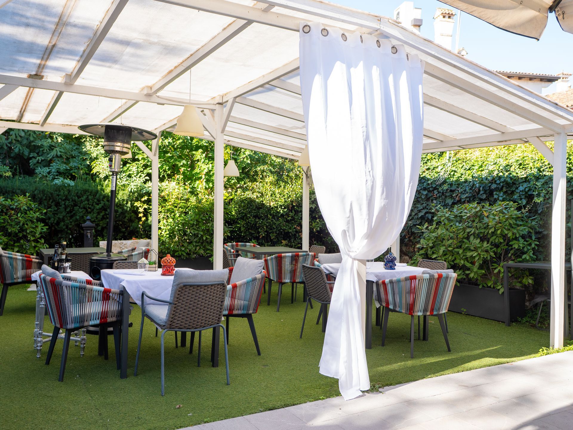 Zona pranzo all'aperto sotto una tettoia bianca con tavoli, sedie e tende bianche. Verde sullo sfondo.