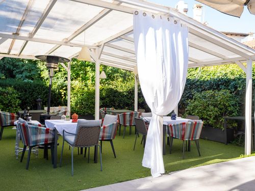 Zona pranzo all'aperto sotto una tettoia bianca con tavoli, sedie e tende bianche. Verde sullo sfondo.