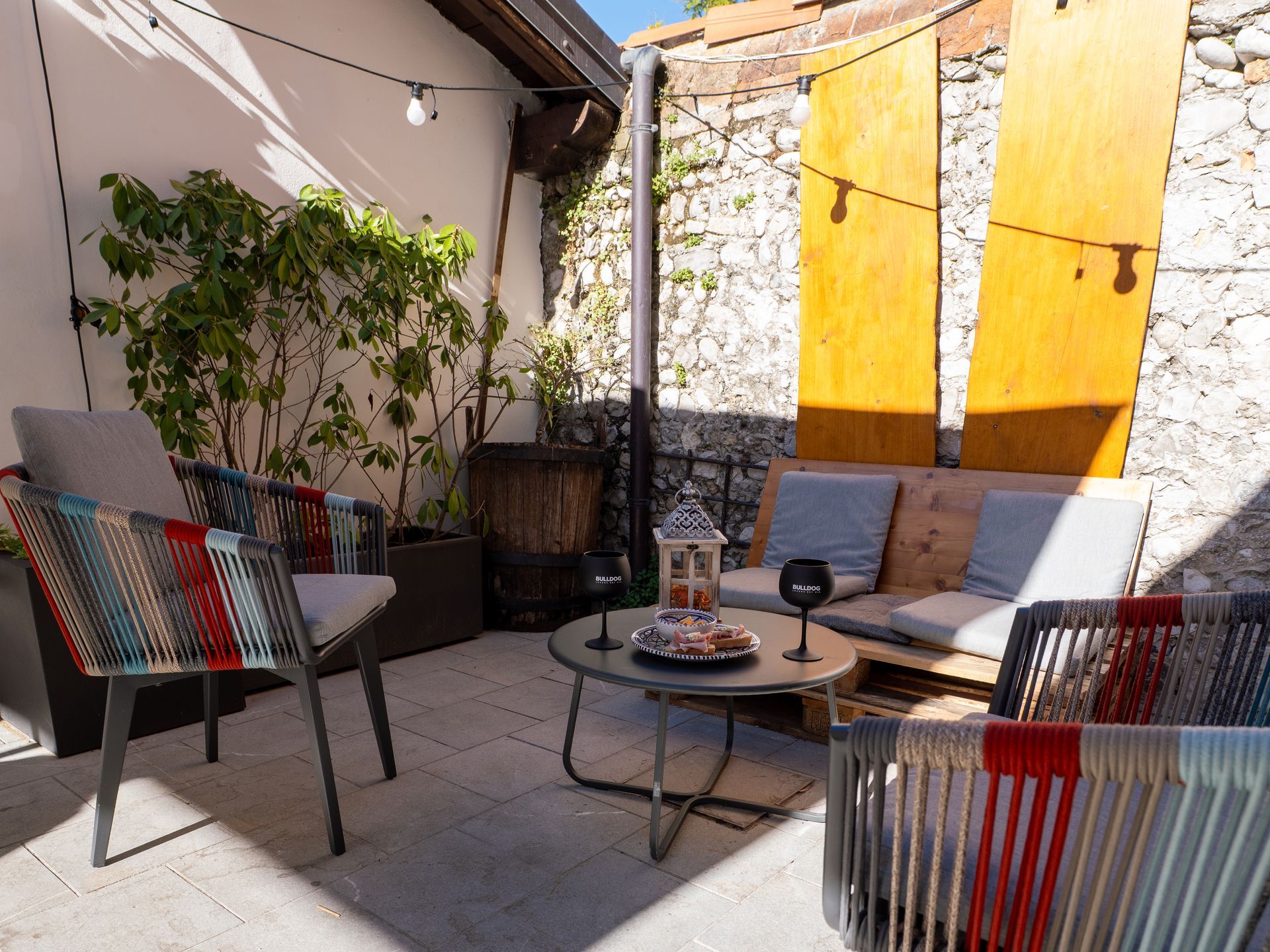 Patio esterno con mobili grigi, un tavolino con uno spuntino e luci a filo contro i muri in pietra.