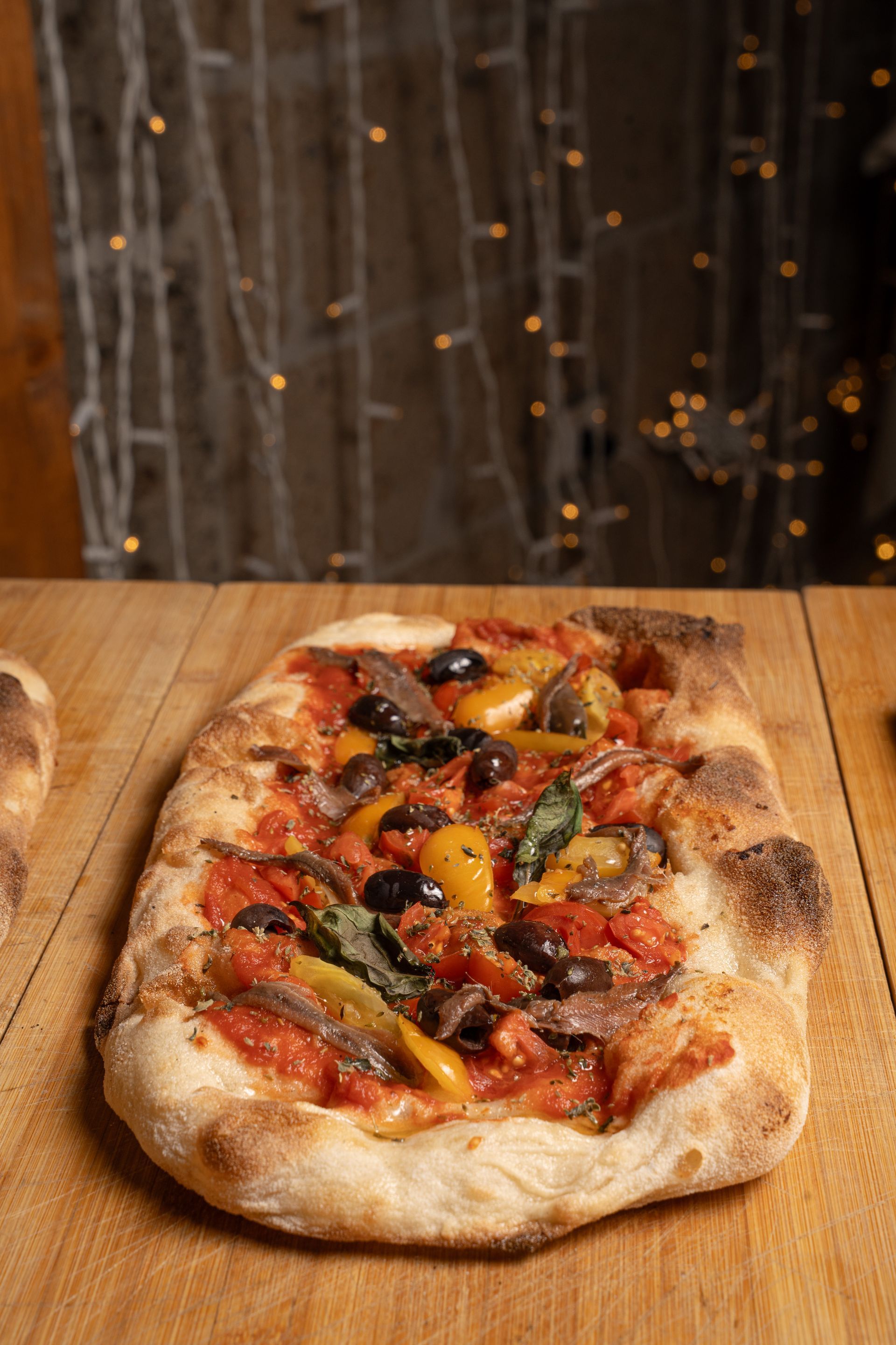 Pizza di focaccia con salsa di pomodoro, olive, acciughe e peperoni gialli su una superficie di legno.
