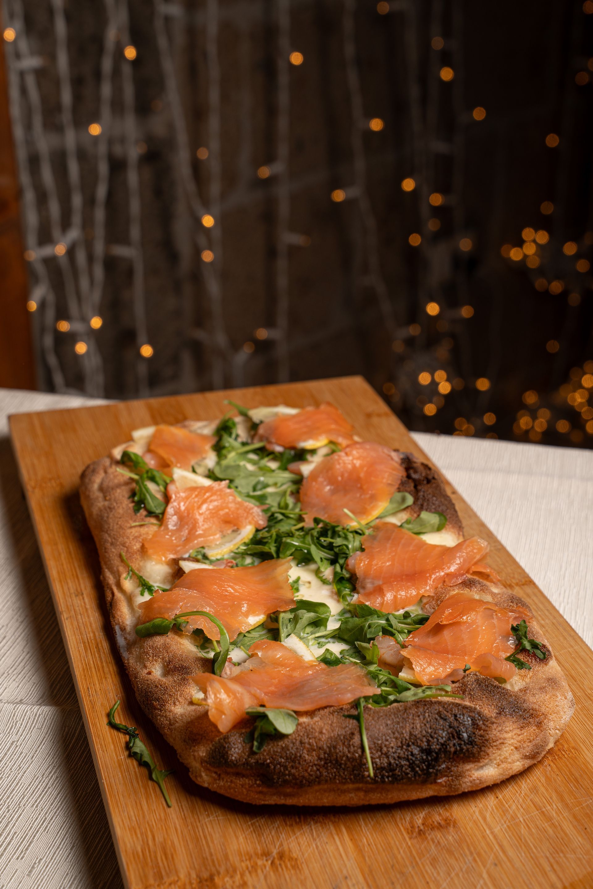 Pizza piatta con salmone e rucola su un tagliere di legno; lucine sullo sfondo.