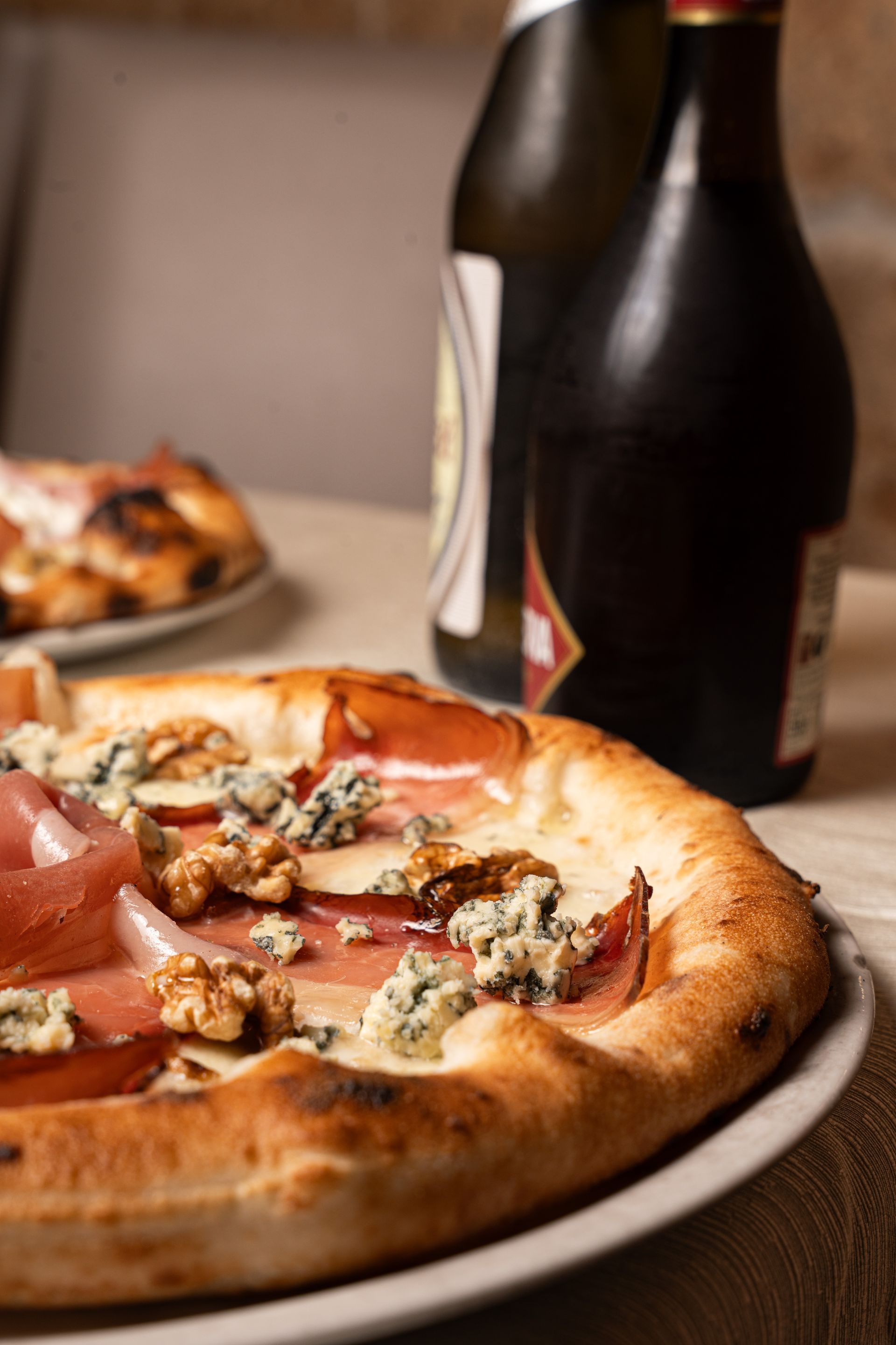 Pizza con prosciutto, noci e gorgonzola, con bottiglie di vino sullo sfondo.