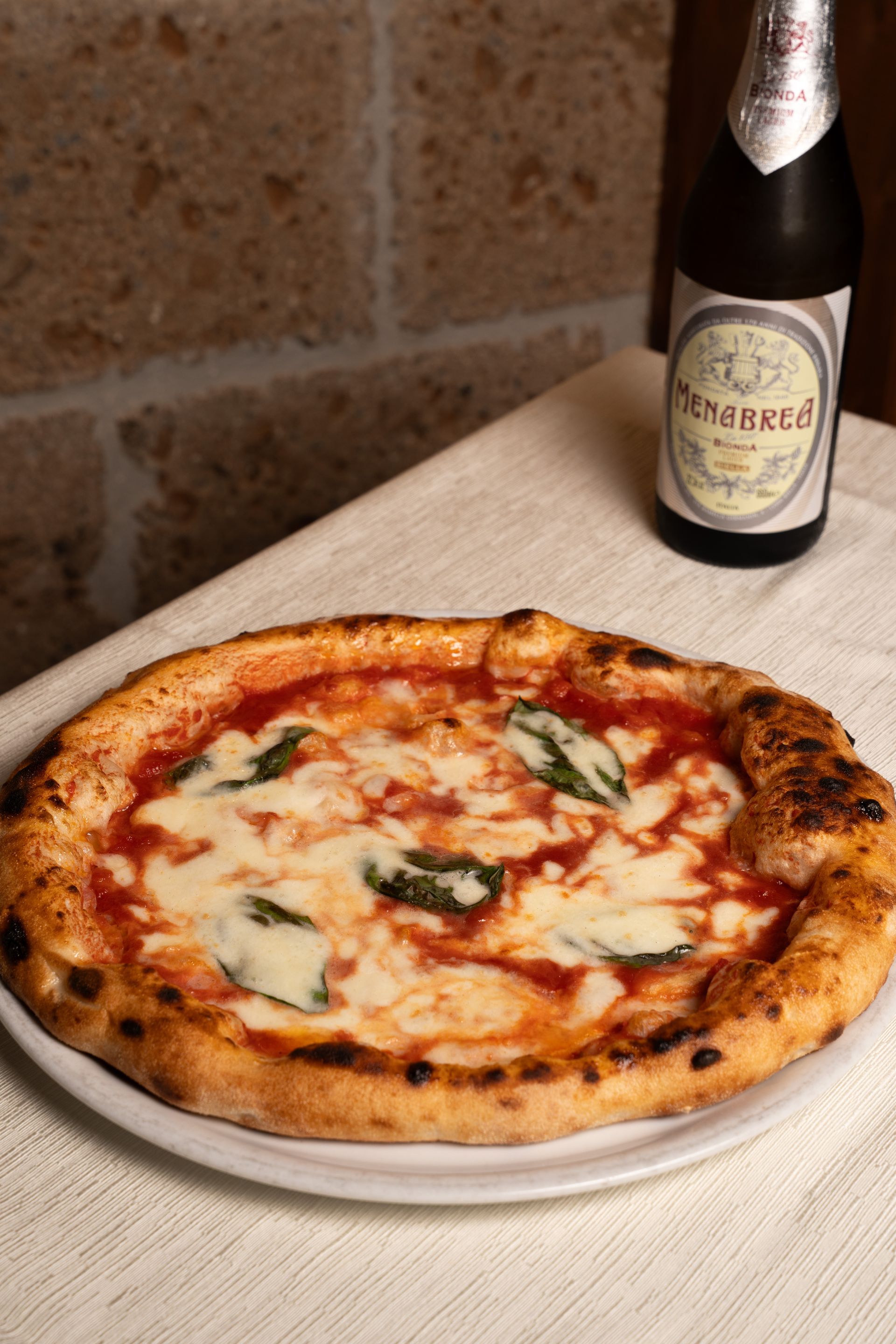 Pizza su un piatto bianco con una bottiglia di spumante su un tavolo con un muro di mattoni sullo sfondo.