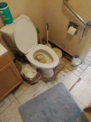 Toilet in a neglected bathroom. Toilet lid up, debris scattered on floor. A blue bath mat in foreground.