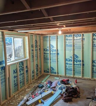 Interior view of a room under construction with insulation panels and a window. Green and wood tones dominate the scene.