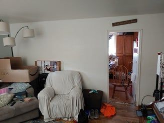 Cluttered living room with a covered chair, doorway to another room, and boxes/furniture.