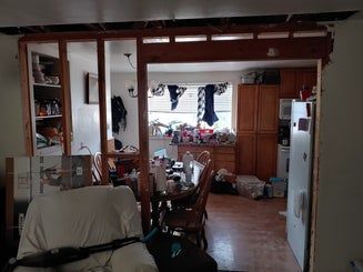 Interior view of a partially demolished wall, revealing a cluttered kitchen and living space.
