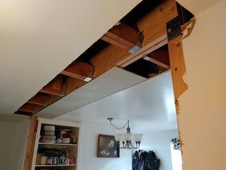Opening in a wall reveals exposed wooden beams and drywall. A dining room with a chandelier is visible.