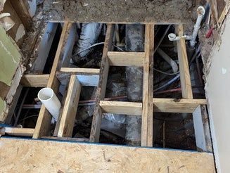 Wooden floor joists with exposed pipes and wiring under a subfloor.