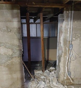 Opening in a concrete wall with damaged edges, wooden supports, debris, and wiring visible.