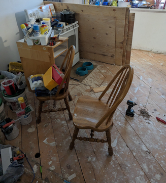 Two wooden chairs on a messy wood floor. Renovation debris surrounds an oven.