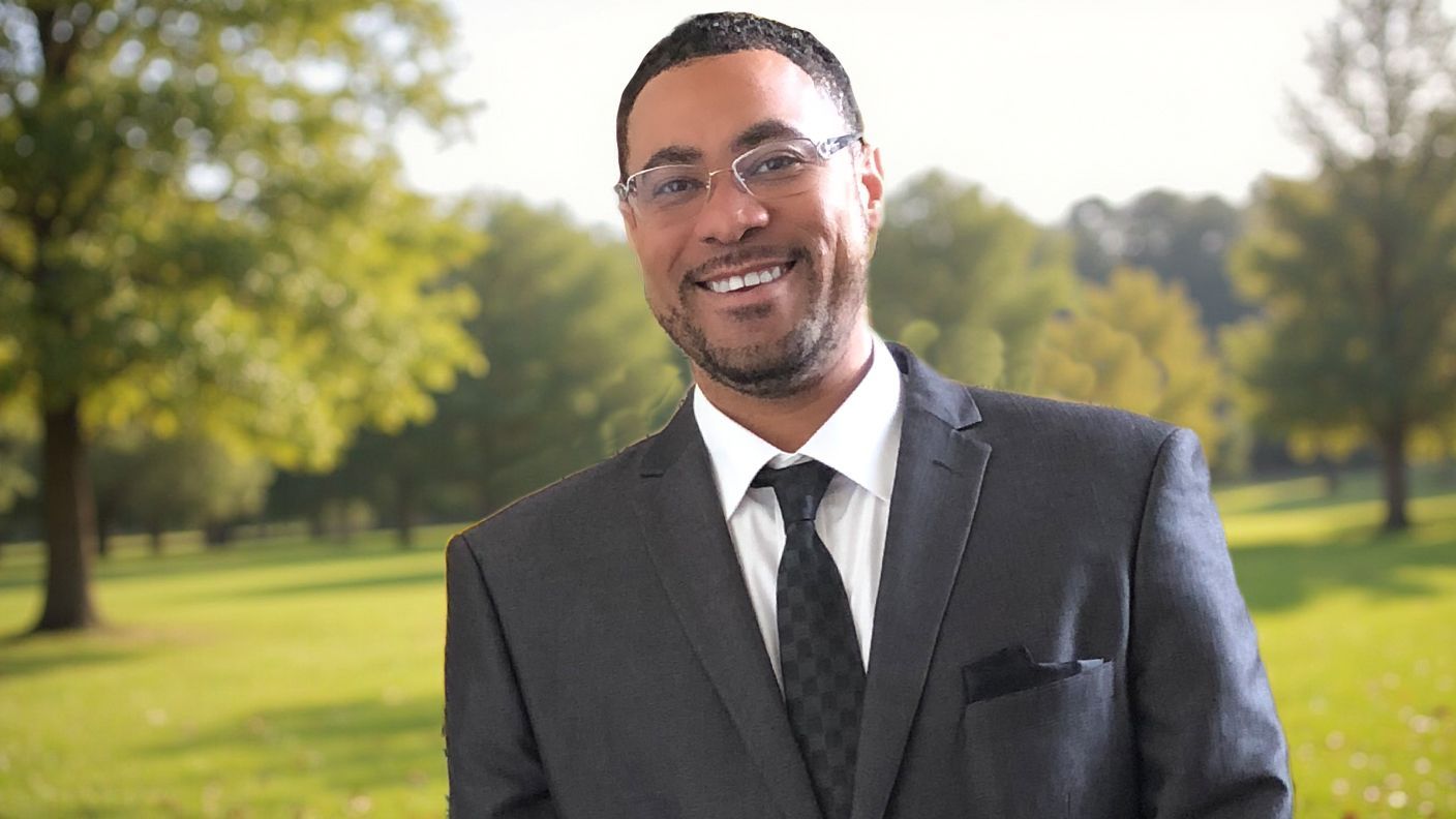 Man in a suit smiles outdoors, green trees and grass in the background.