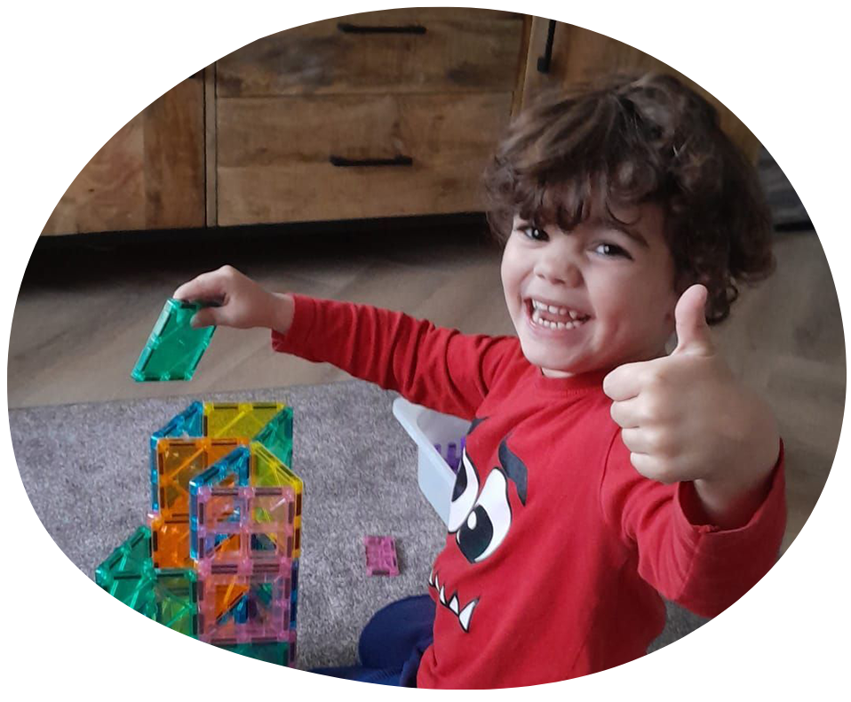 Drie lachende kinderen liggen op een oppervlak, één met een potlood in de hand, omringd door kleurrijke voorwerpen.