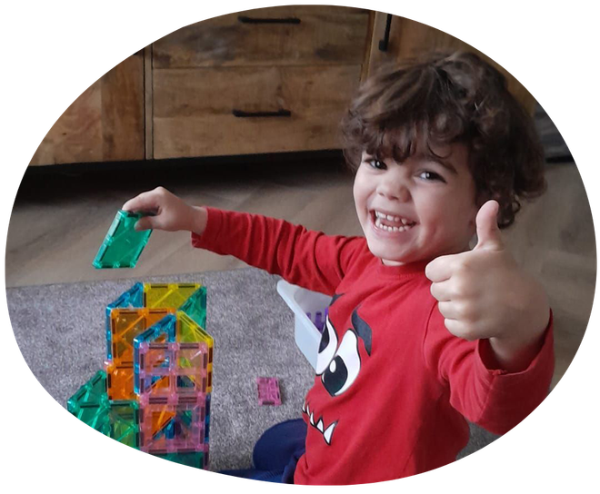 Drie lachende kinderen liggen op een oppervlak, één met een potlood in de hand, omringd door kleurrijke voorwerpen.