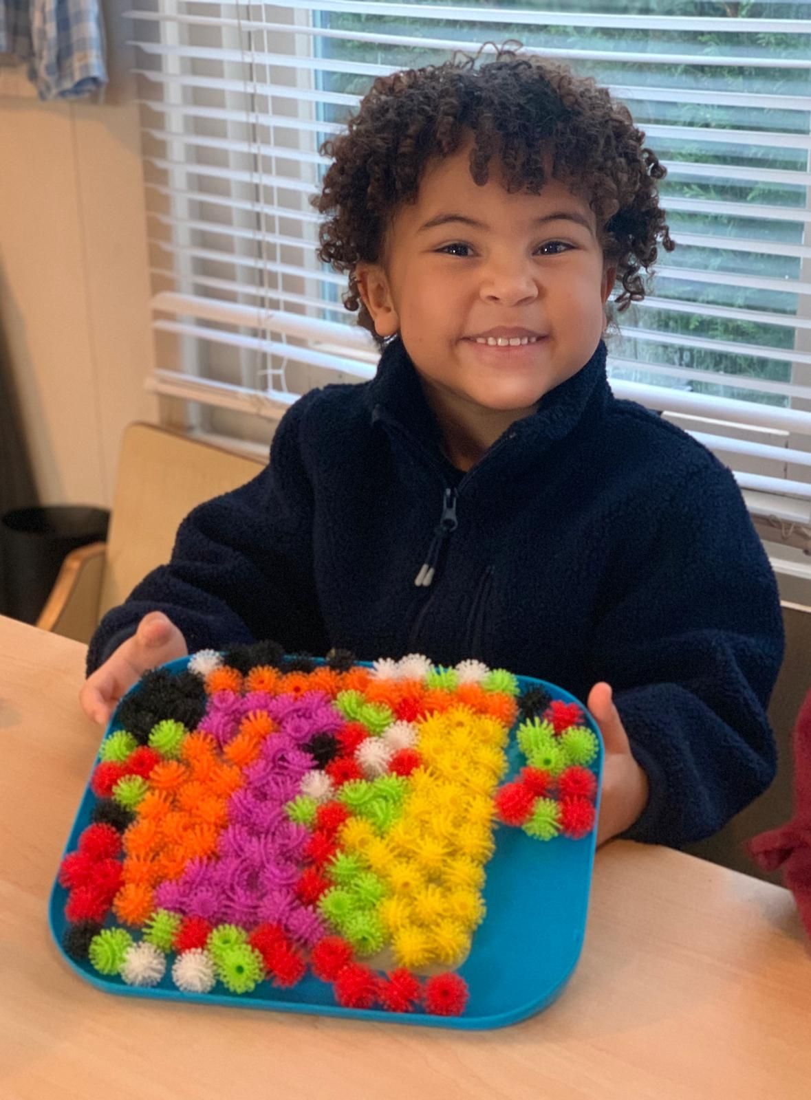 Een kind in een spijkerbroek en een vestje houdt ovenwanten vast en staat bij een speelkeukentje.