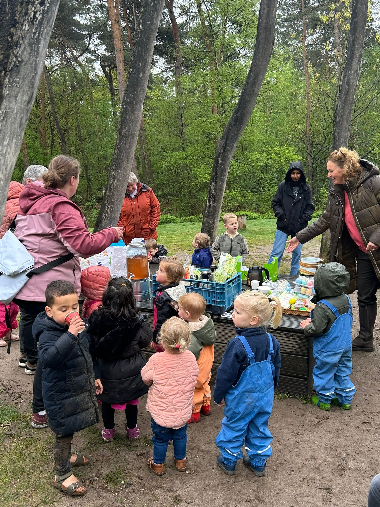 Een kind geeft buiten de planten water in een metalen kan, naast een kleine rode driewieler.
