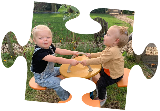 Een jong kind in een roze jasje zit in een rood wagentje, getrokken door een jongen op een gele speelgoedtractor.