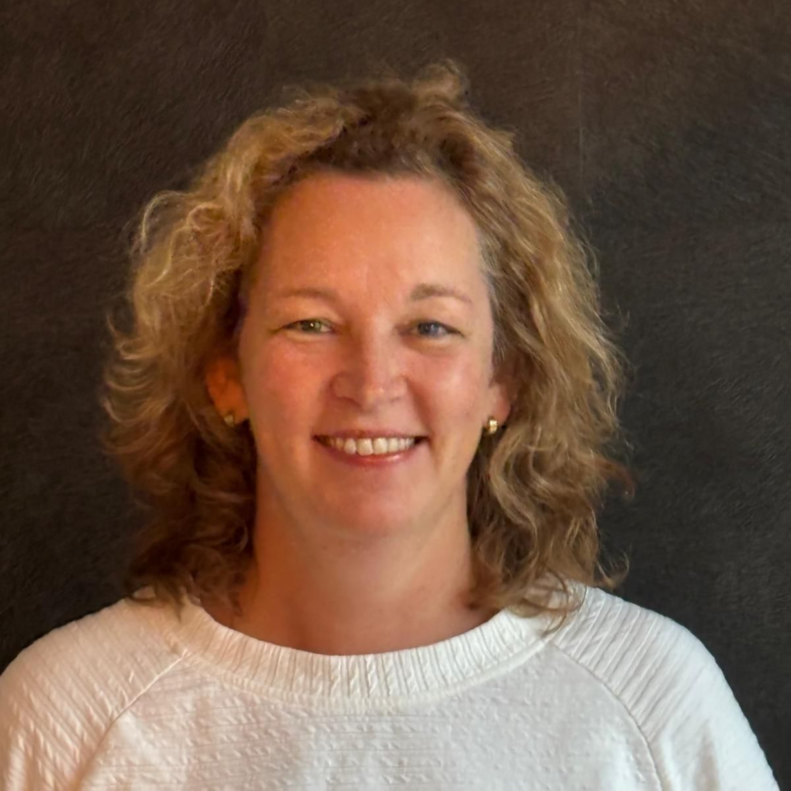 Vrouw met krullend blond haar lacht naar de camera, gekleed in een witte trui.