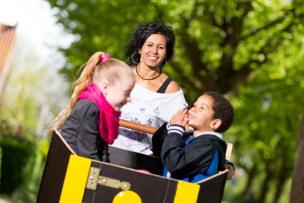 Lachende vrouw met twee kinderen in een kartonnen doos buiten, zonnige dag.