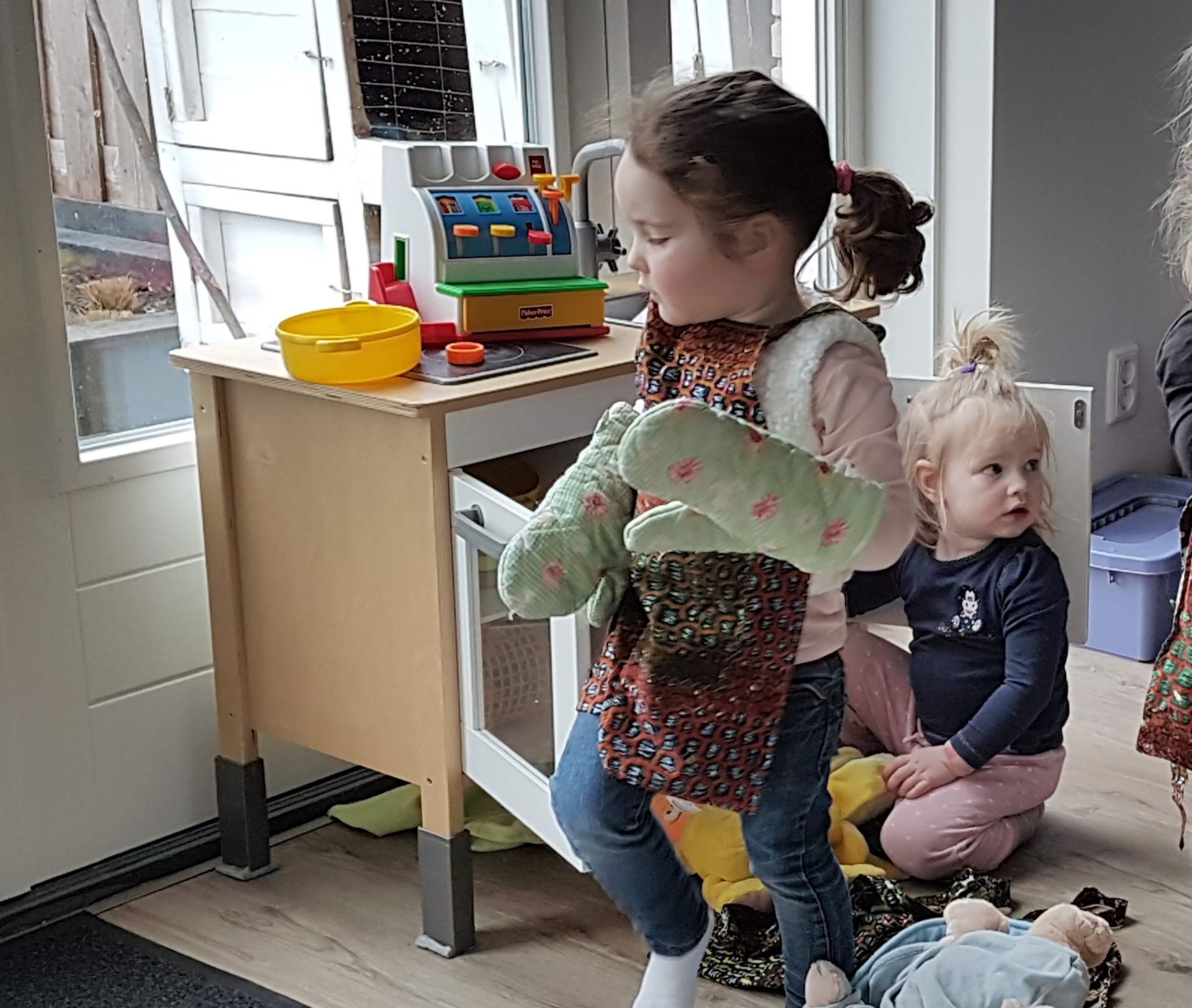 Een kind in een spijkerbroek en een vestje houdt ovenwanten vast en staat bij een speelkeukentje.