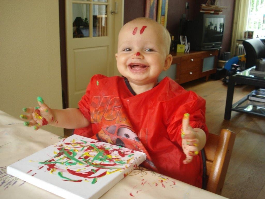 Blije baby met verf op gezicht en handen, schilderend op een doek aan tafel. Rood schortje.